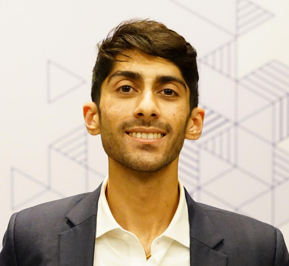Headshot of Prathm Juneja in a navy blue suit and white shirt against an unfocused geometric background